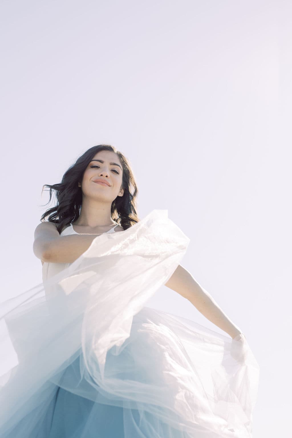 A bride in a pale blue tulle gown smiling as her skirt catches the wind