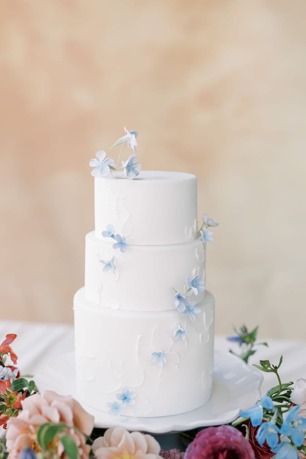 A three tier white wedding cake with pale blue sugar flowers beside a peach and berry bouquet