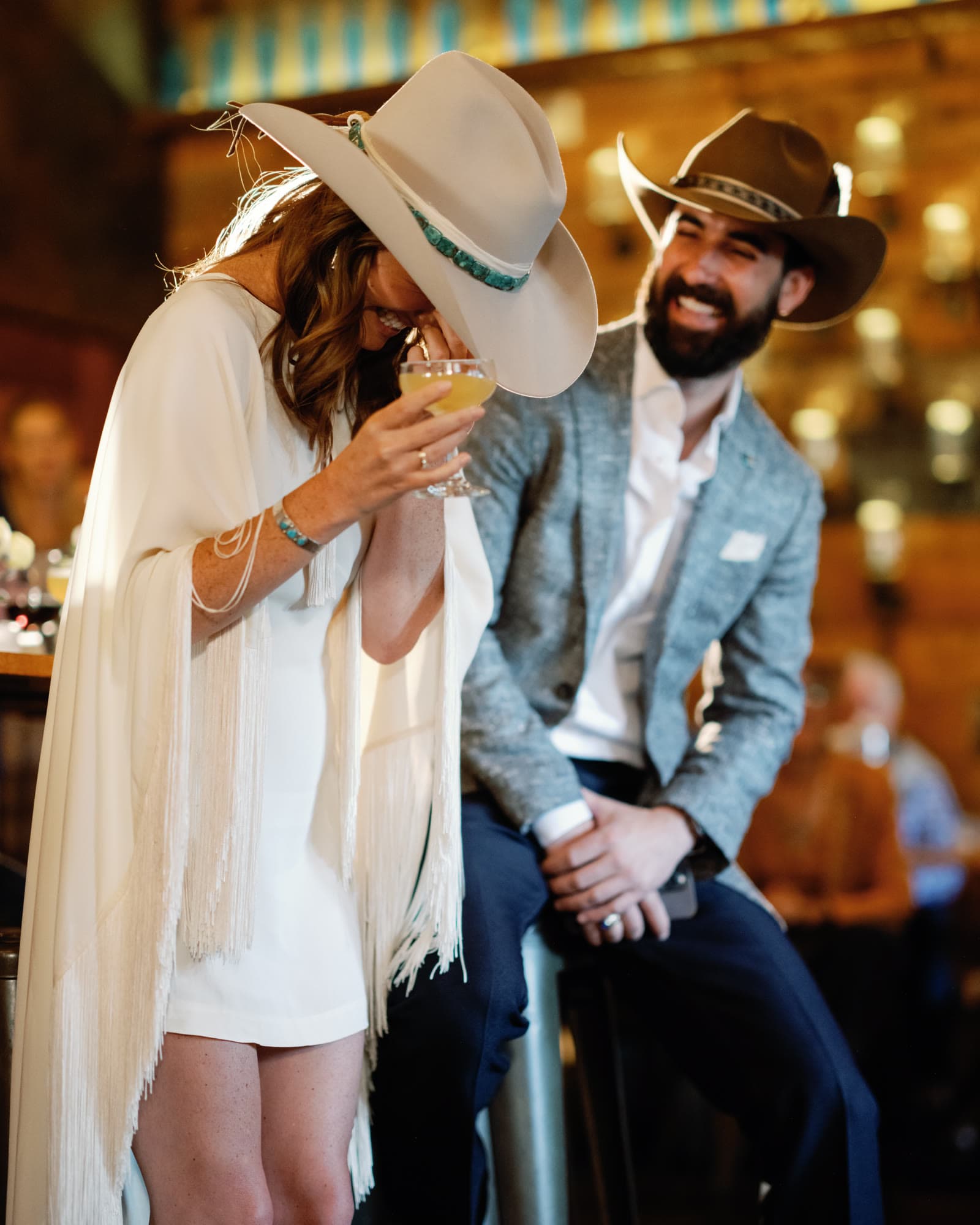 A bride in a fringed cape and cowboy hat laughing over a cocktail with her groom at the reception bar