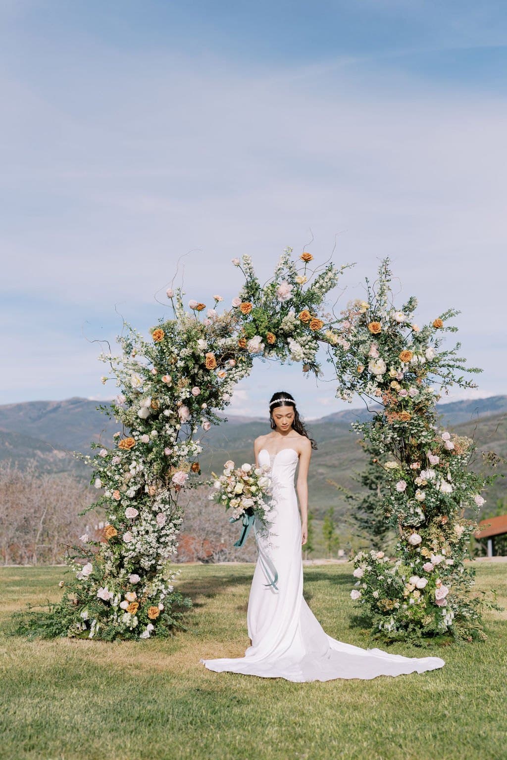 A bride beneath a peach and white floral ceremony arch with the Park City mountains behind her