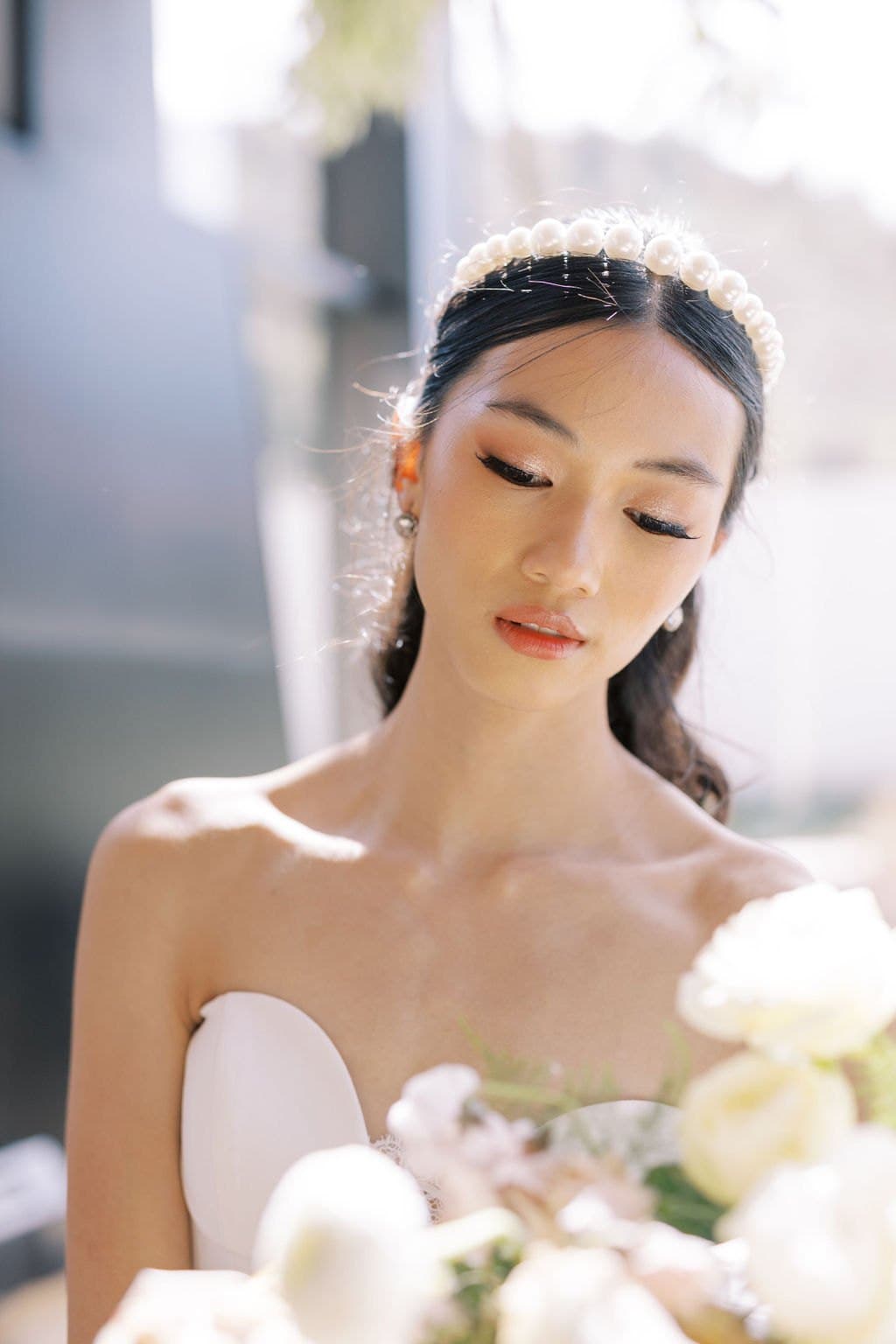A bride in a pearl headband looking down at her bouquet in soft light