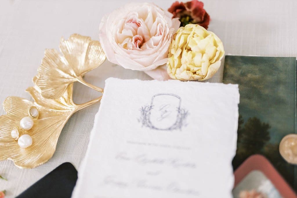 A deckled edge wedding invitation with a monogram crest beside pearl earrings and garden roses