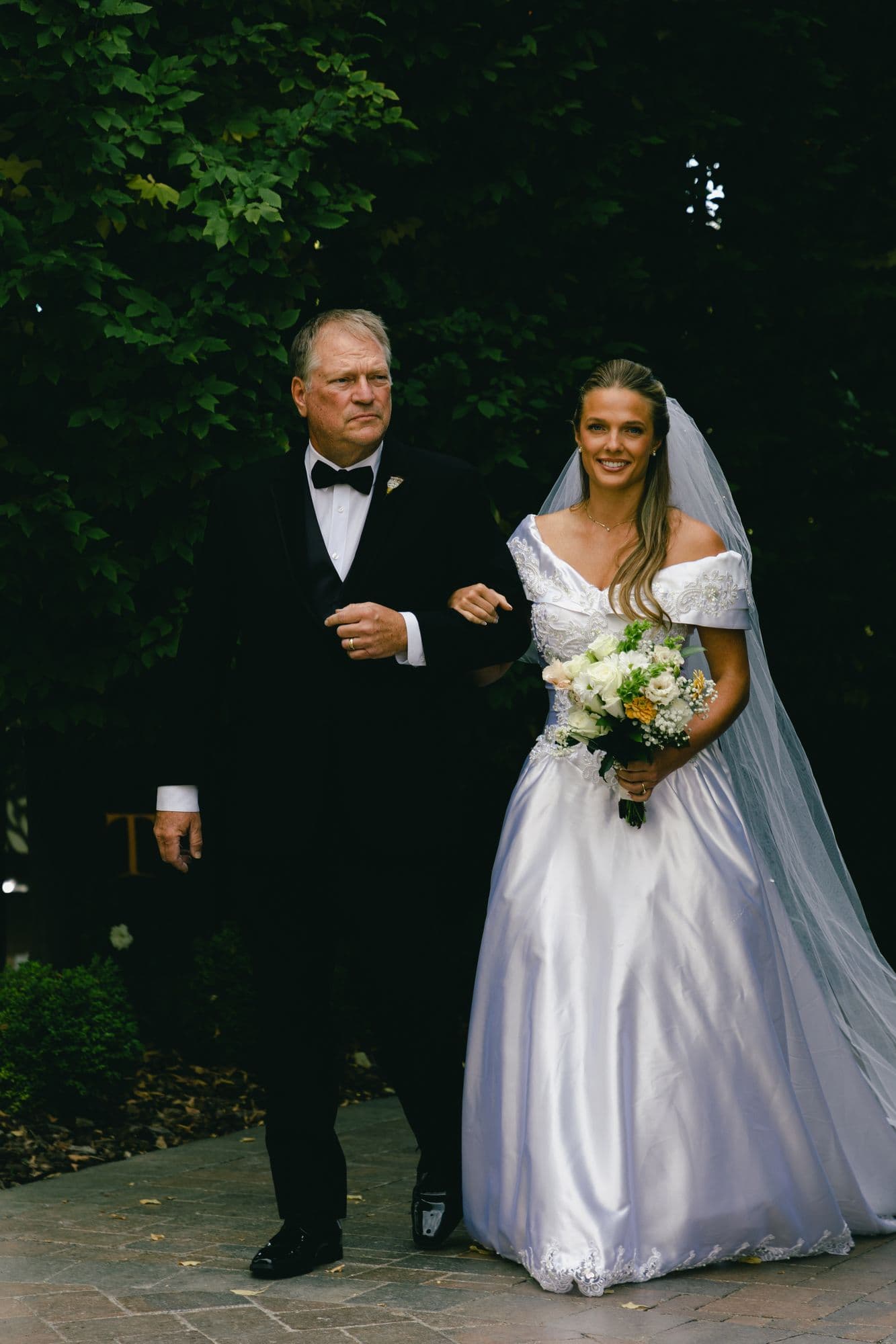 Marita walking up the brick aisle on her father’s arm, veil down, white bouquet in her hand