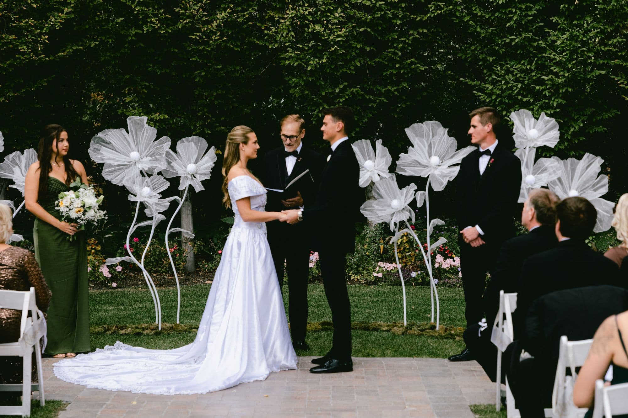Kyle and Marita holding hands during their ceremony at Bellissimo Gardens, oversized white paper flowers on either side of them and guests seated in white chairs