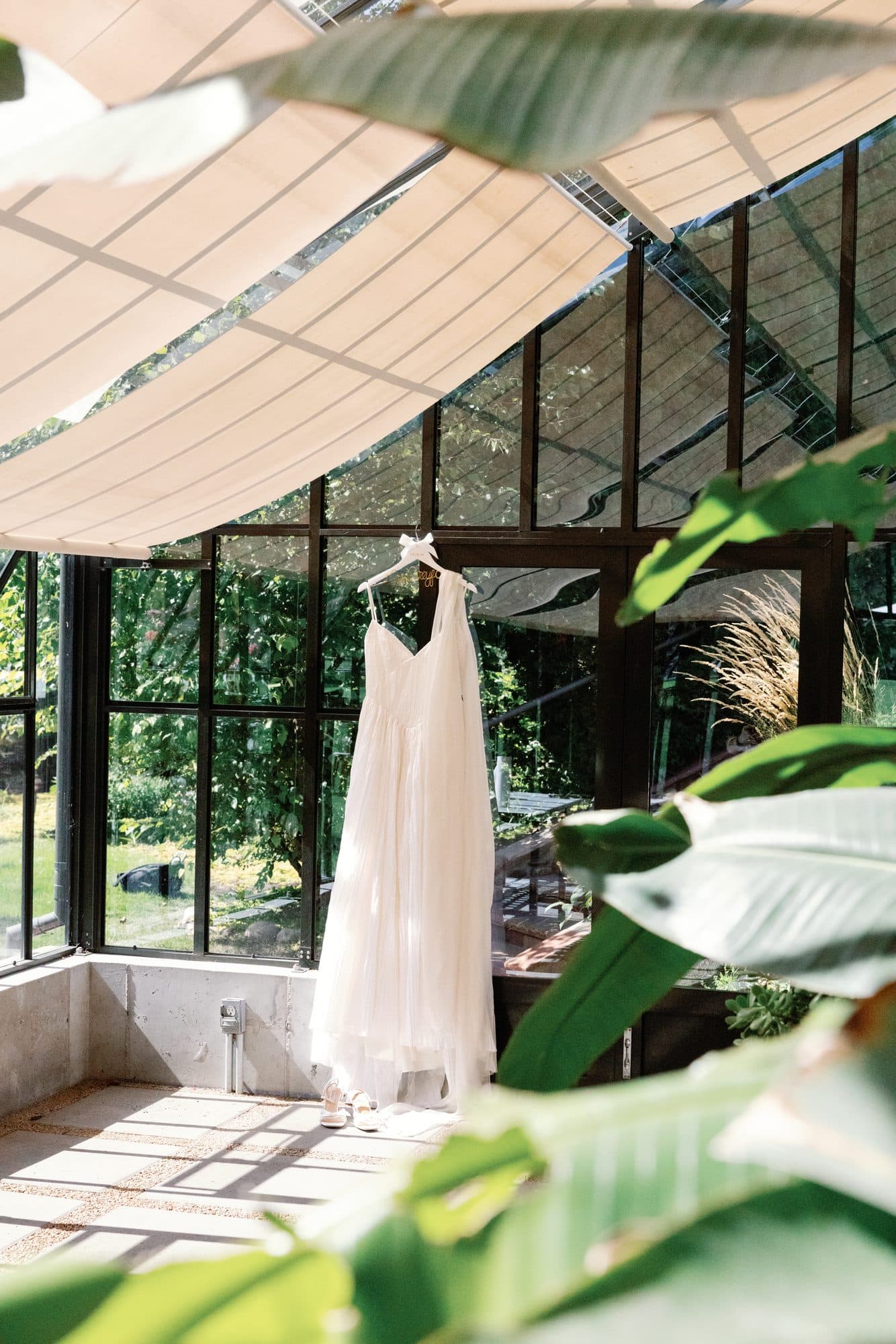 Lauren’s wedding dress hanging in a glass greenhouse, banana leaves and morning light in front of it