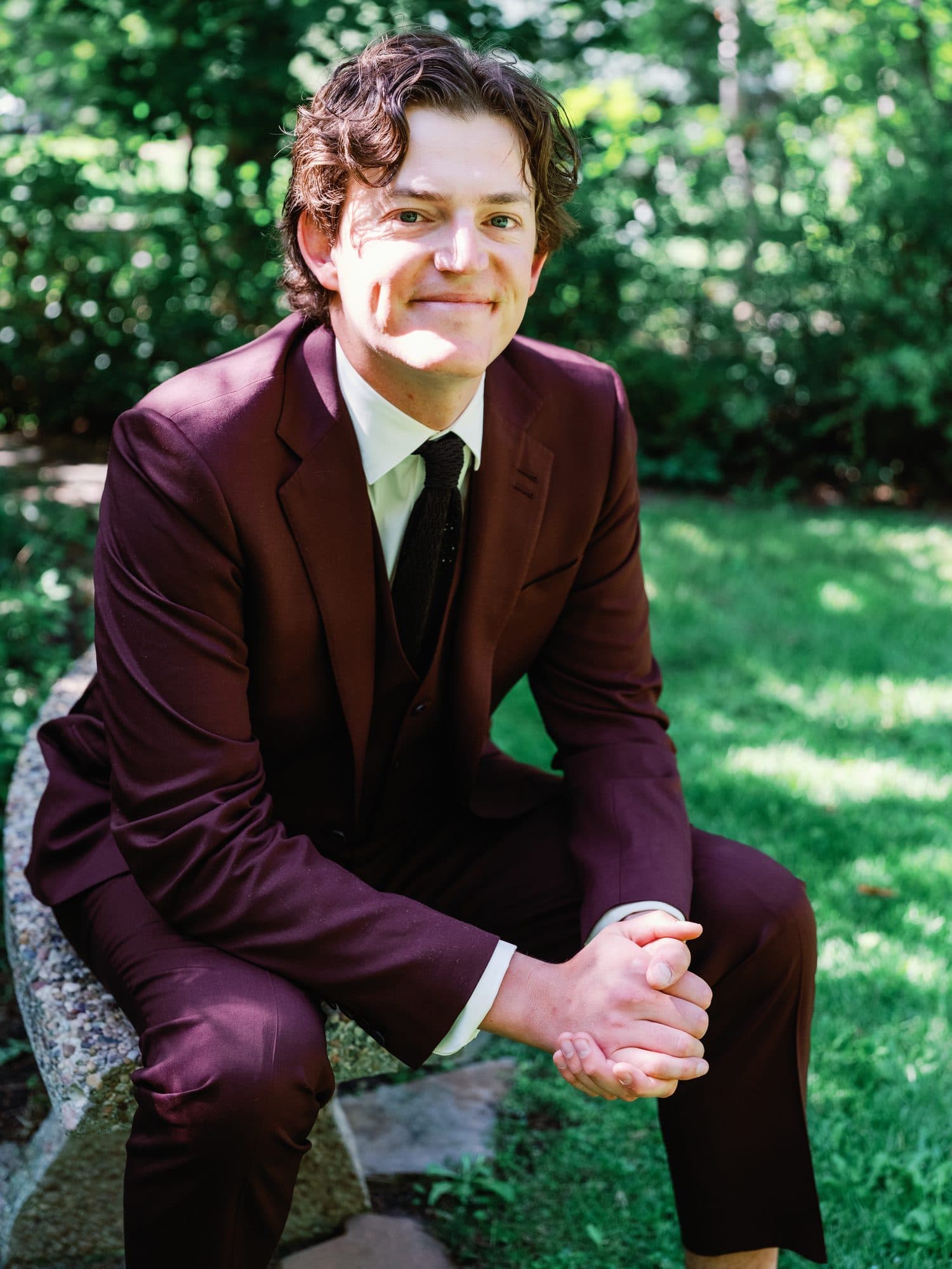 Jake in his burgundy suit, sitting on a stone wall in the garden before the ceremony