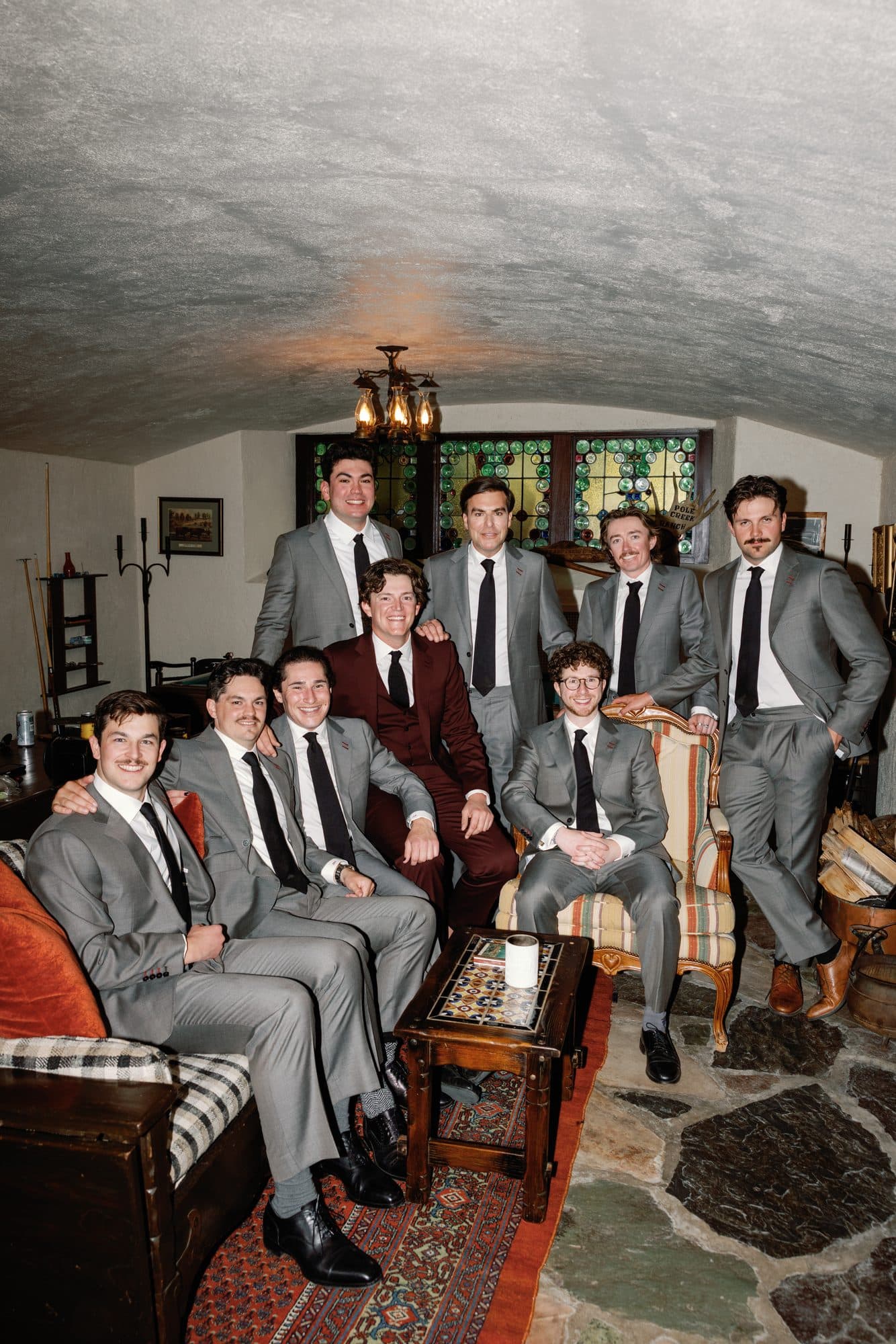 Jake in burgundy with his groomsmen in grey suits, gathered in a stone-floored room