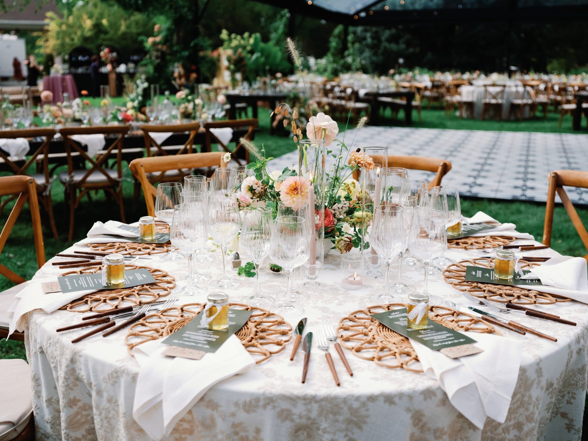 A round reception table set on the lawn with rattan chargers, green menus and a small jar of honey at each place