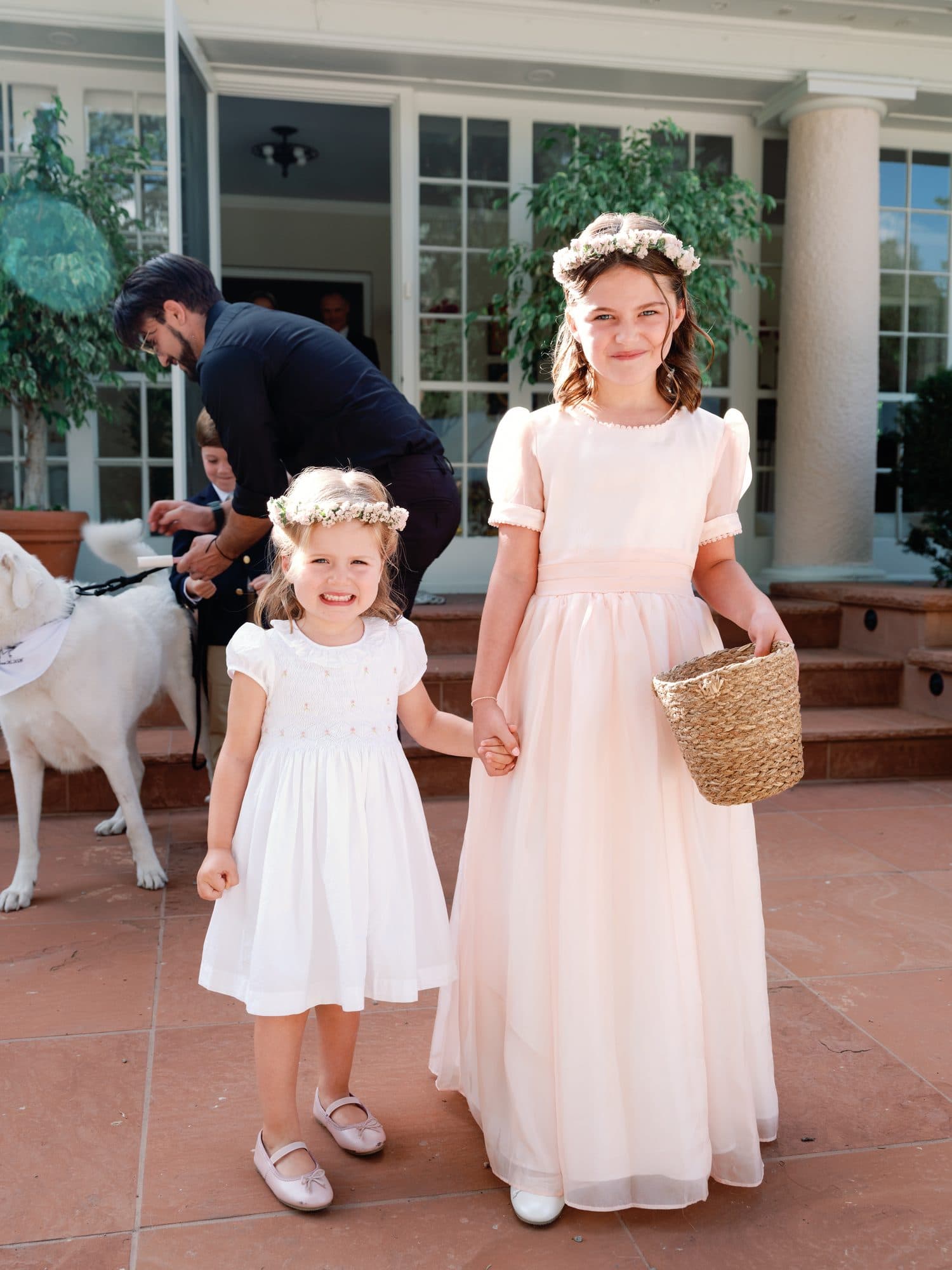 Two flower girls with flower crowns on the steps, a white dog behind them