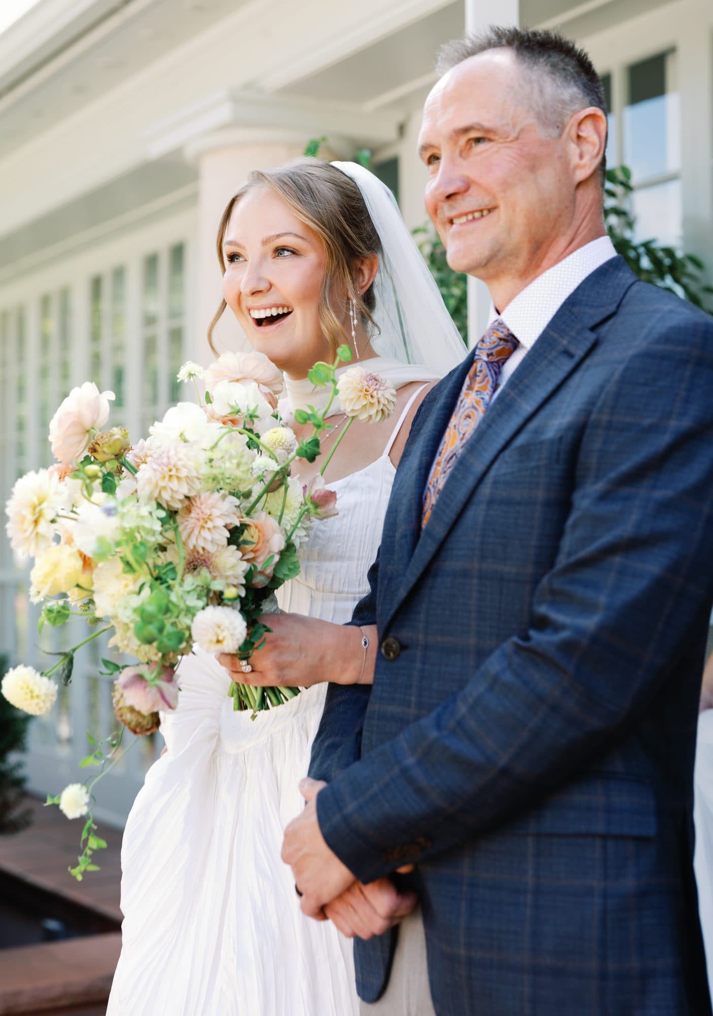 Lauren laughing beside her father on the steps just before the aisle, holding her bouquet of peach and cream dahlias