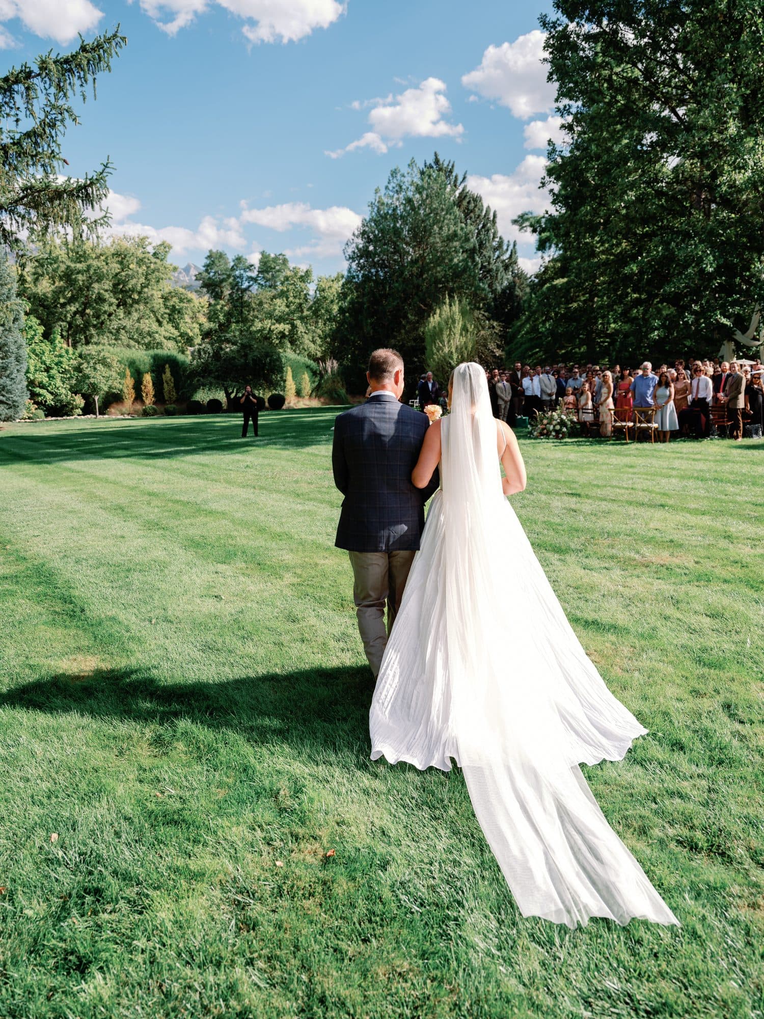 Lauren walking across the lawn toward the ceremony on her father’s arm, her long veil trailing on the grass