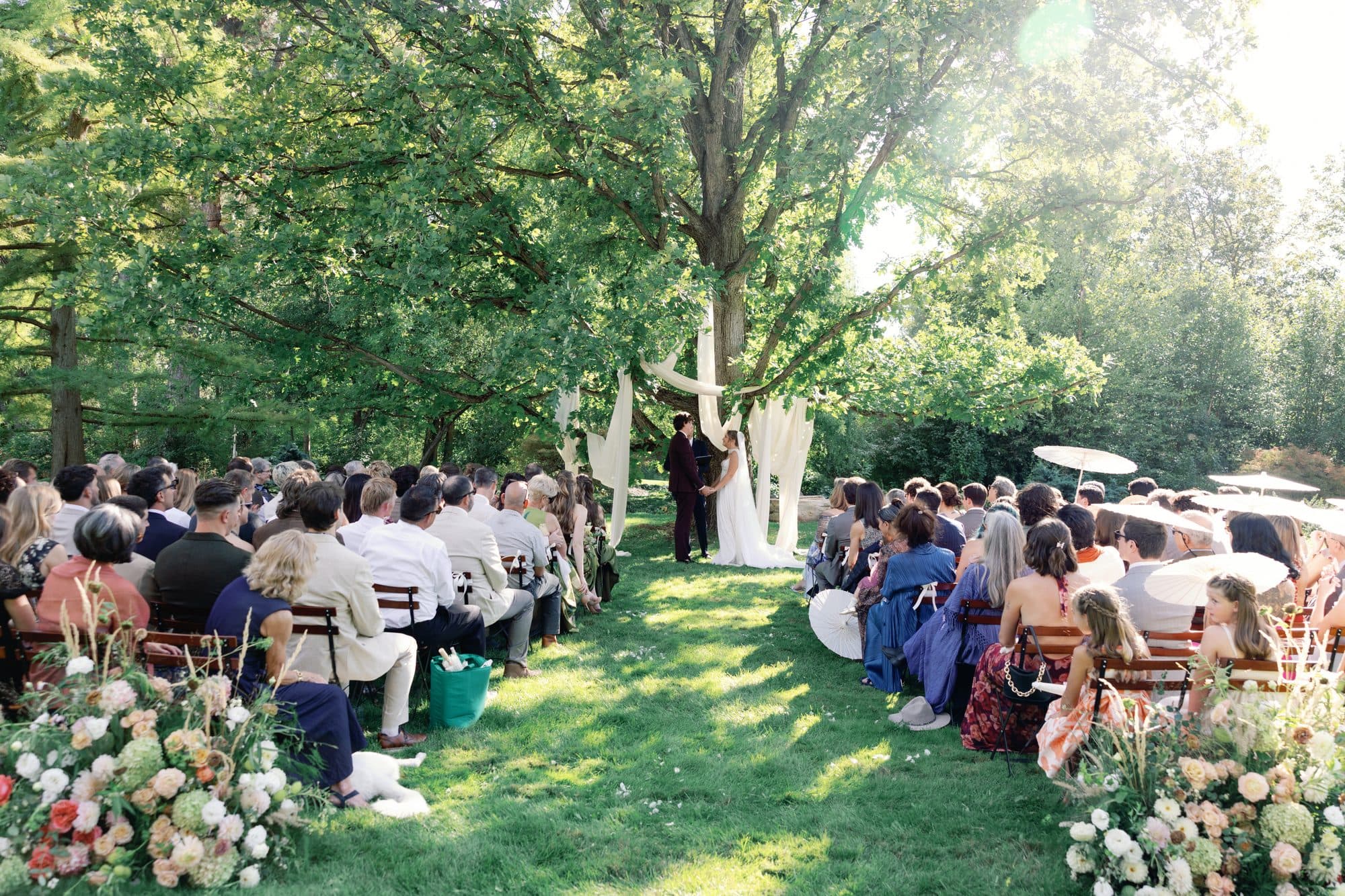 The ceremony from the back of the aisle: guests seated on the lawn between banks of flowers, Lauren and Jake under the old tree draped in white