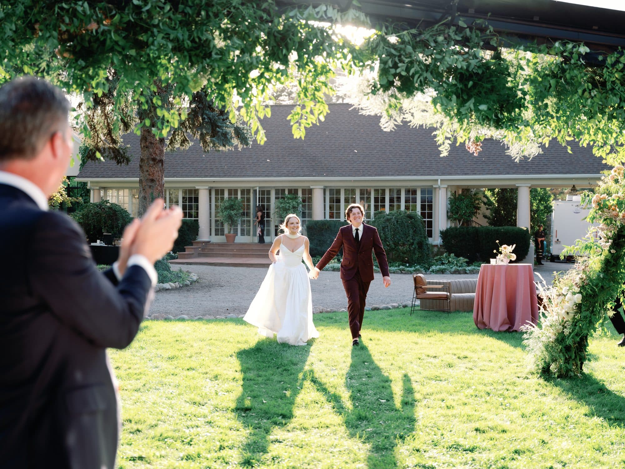 Lauren and Jake making their entrance across the sunlit lawn as a guest applauds in the foreground