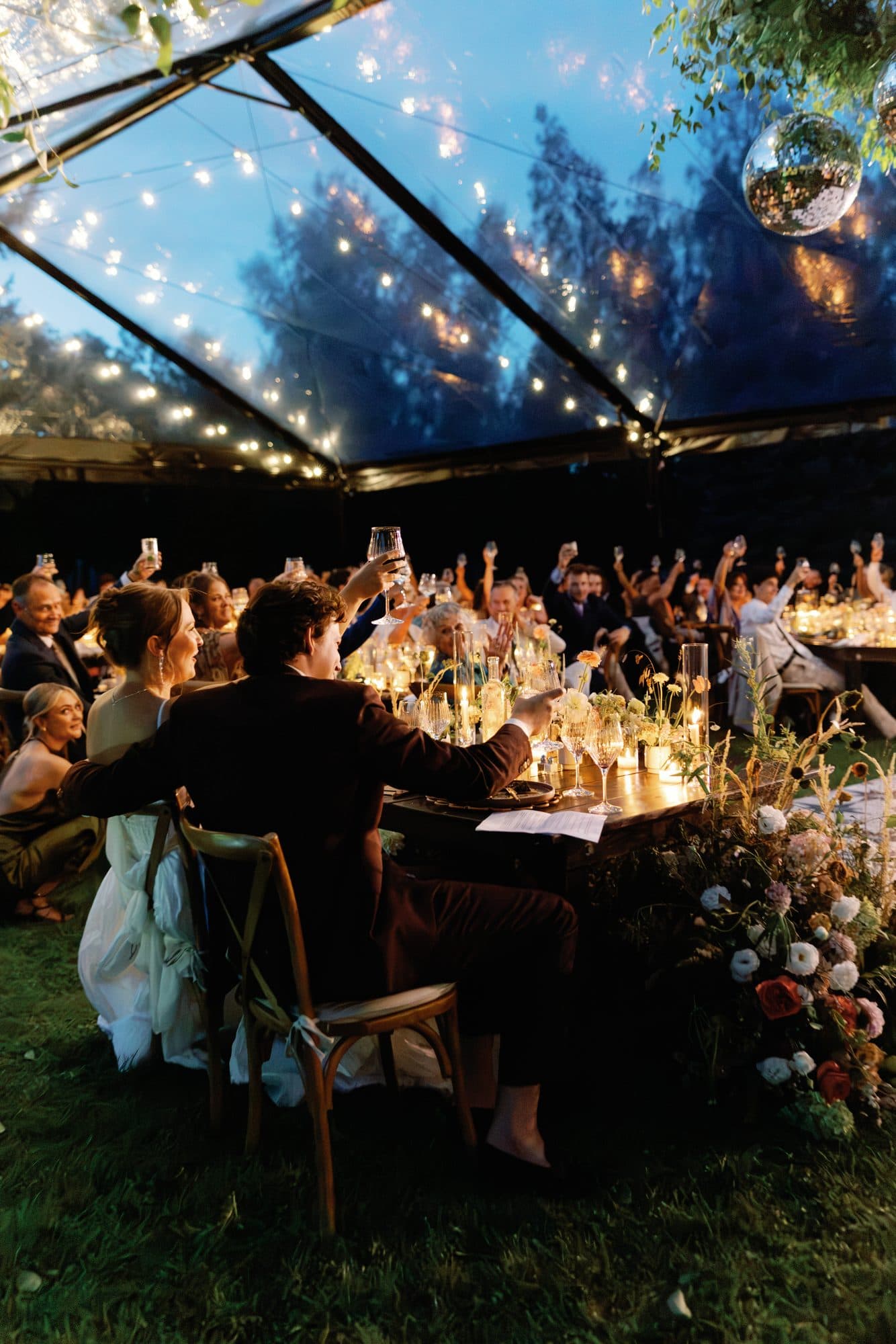 Guests raising their glasses under the string lights of the clear tent at night