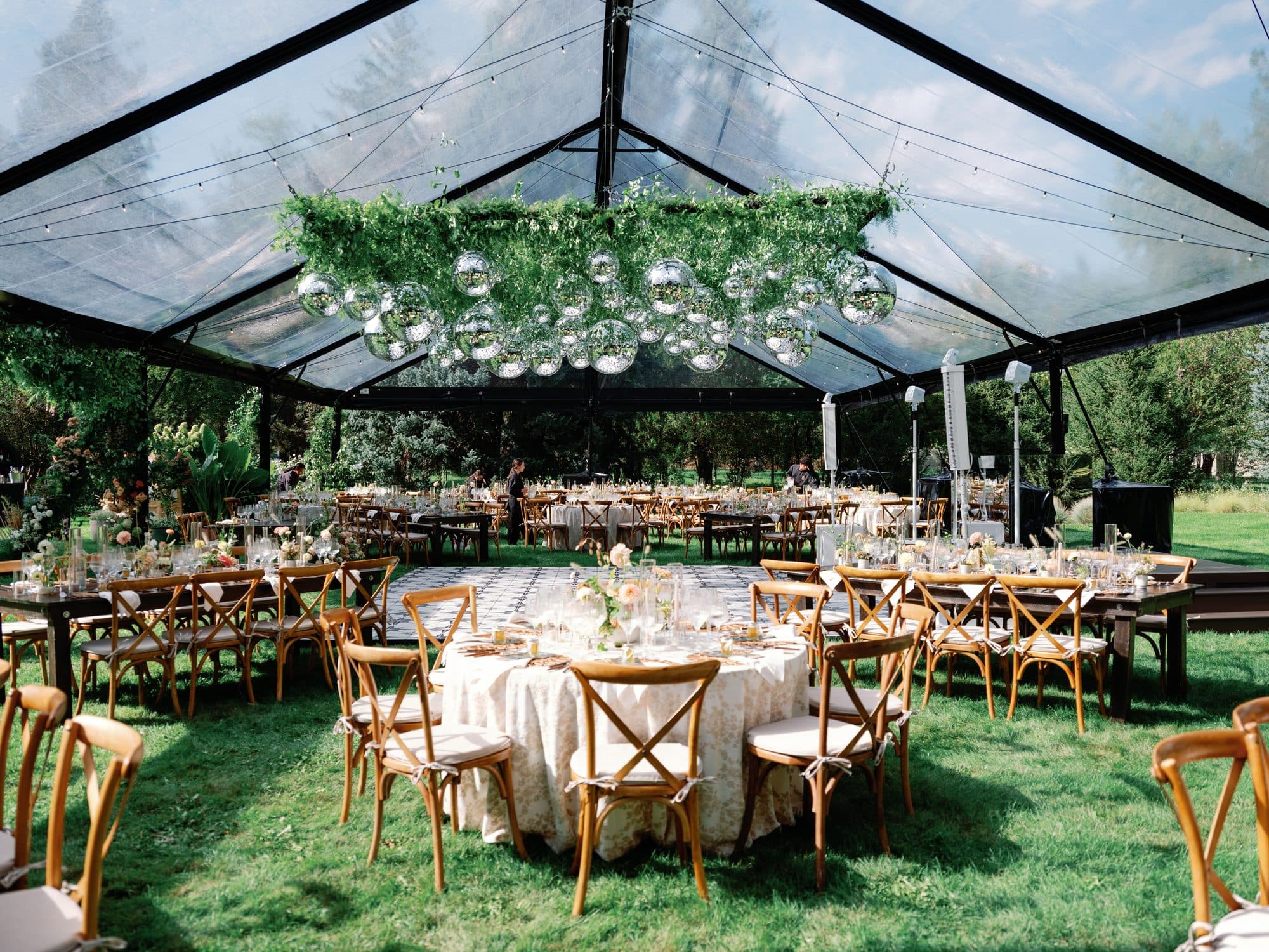 Inside the clear-roofed tent before dinner: round tables in cream linen, crossback chairs, and a hanging garland of greenery and disco balls over the dance floor