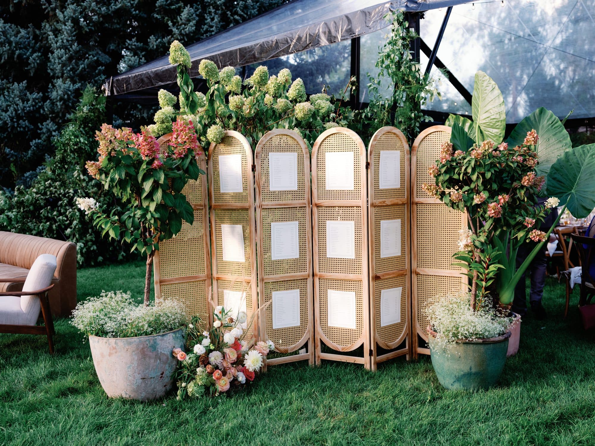 The seating chart set into arched rattan screens on the lawn, flanked by blooming hydrangea trees and elephant-ear leaves