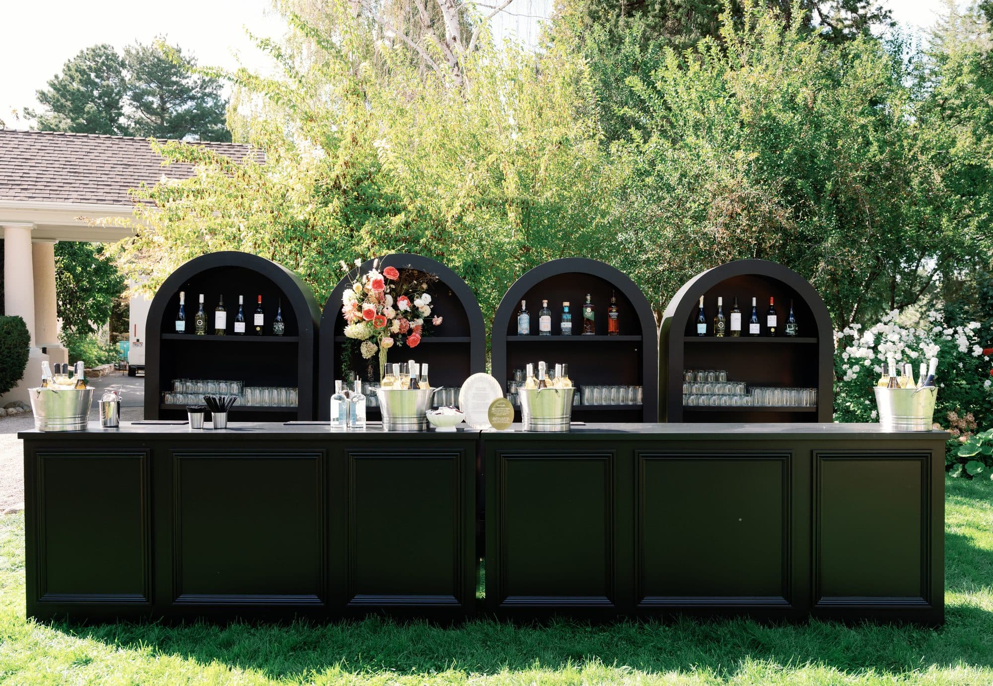 The black bar on the lawn, four arched shelves of bottles behind it and a tall arrangement of garden roses at the center
