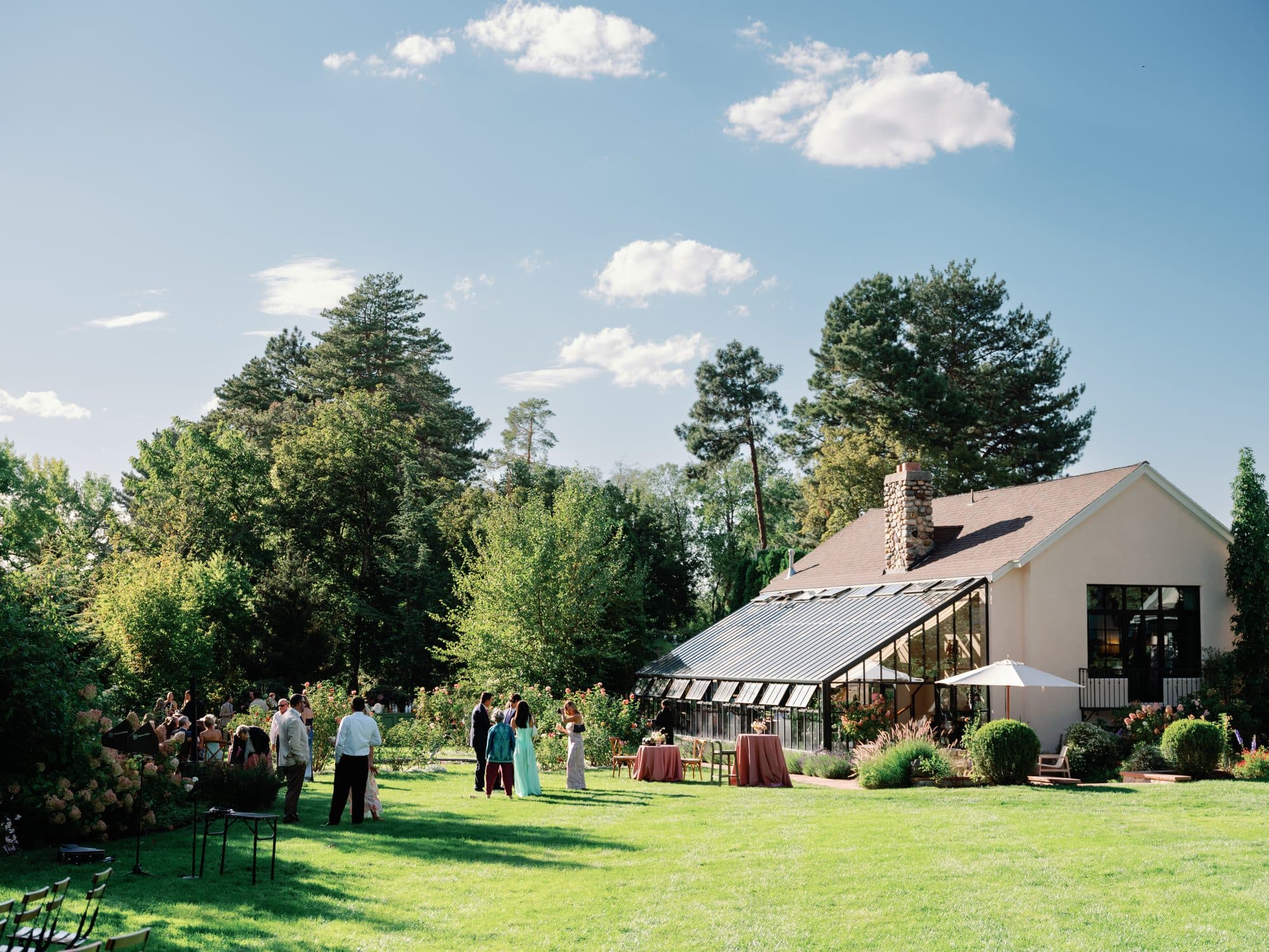 Guests on the lawn at cocktail hour, the glass greenhouse and the house behind them under a blue sky