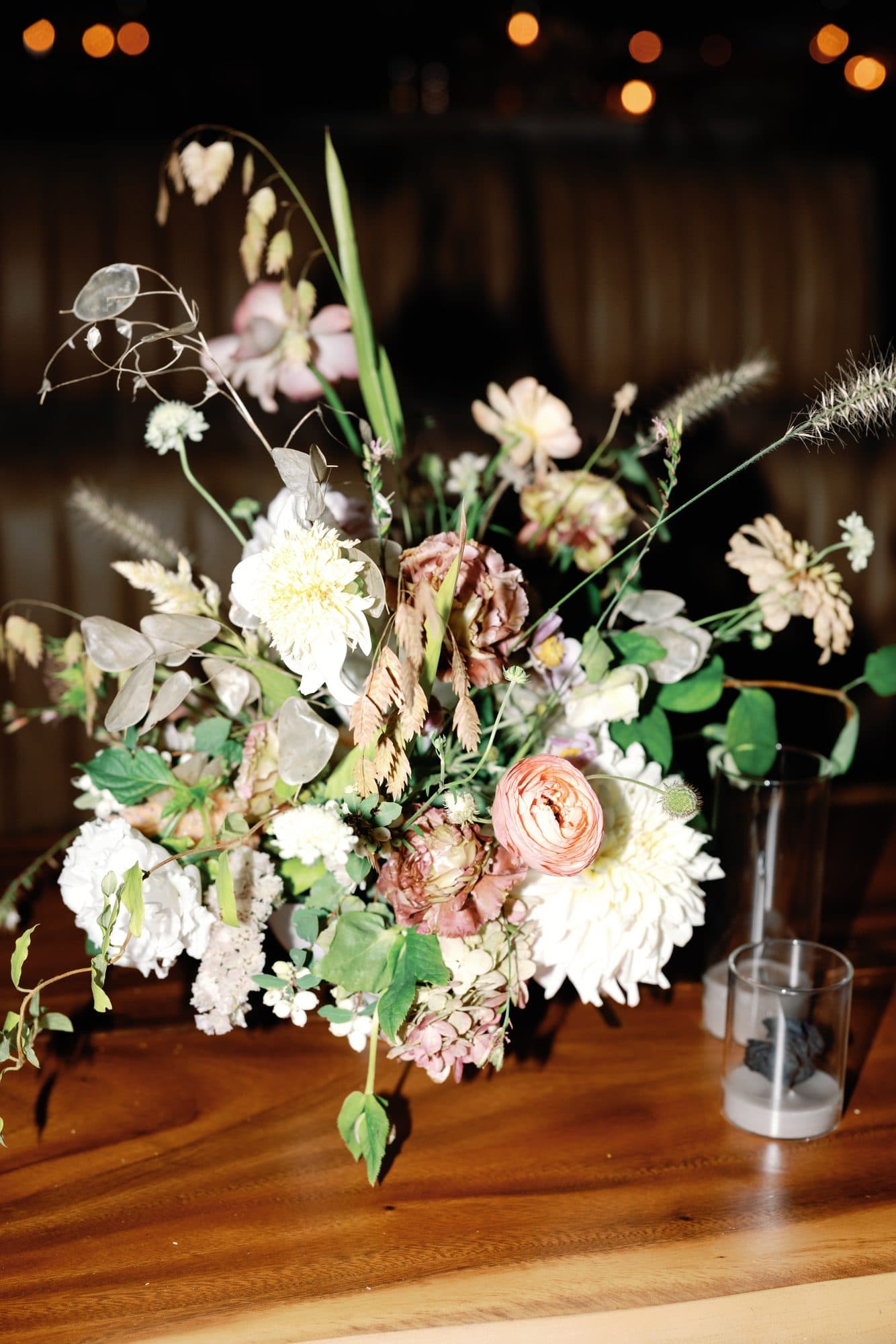 A late-night arrangement of dahlias, ranunculus and dried grasses on a wooden table by candlelight