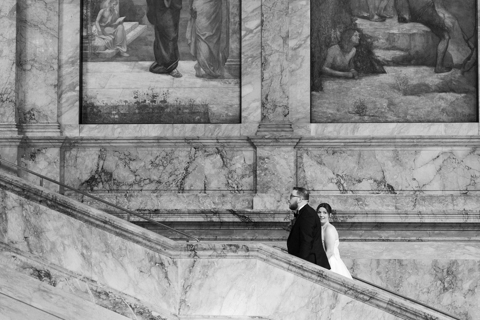 Wide view of Katie and Zach crossing the grand staircase beneath the murals of the Boston Public Library, in black and white