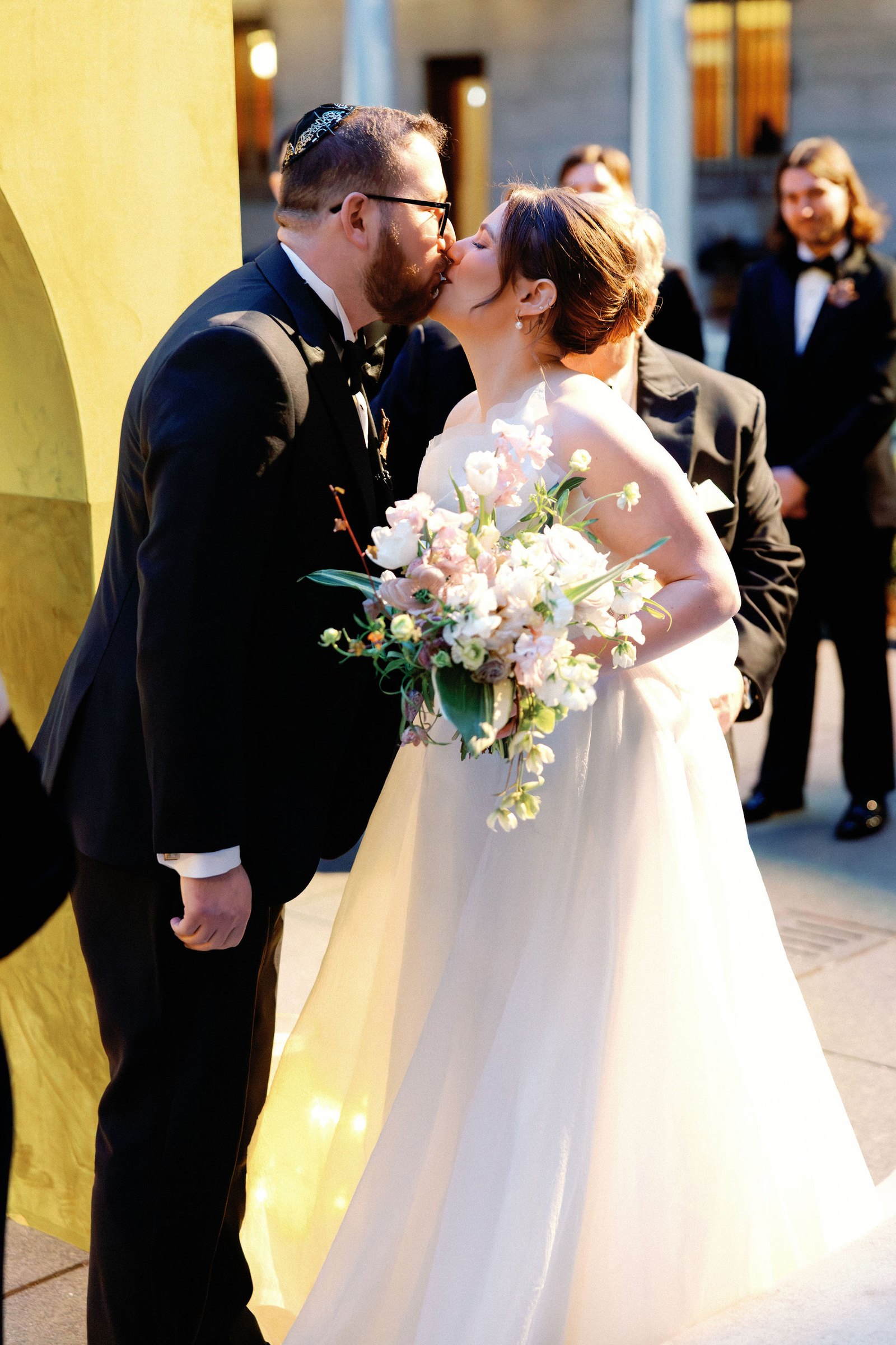 Katie and Zach kiss under the arcade of the Boston Public Library courtyard, her bouquet of pale pink blooms between them