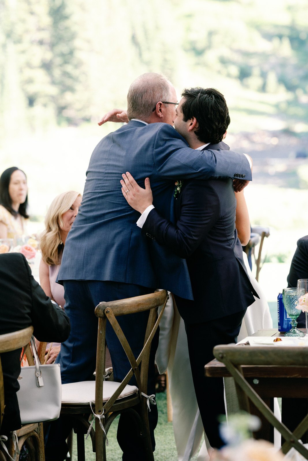 Andrew embraces a guest between the dinner tables, the green mountain behind them