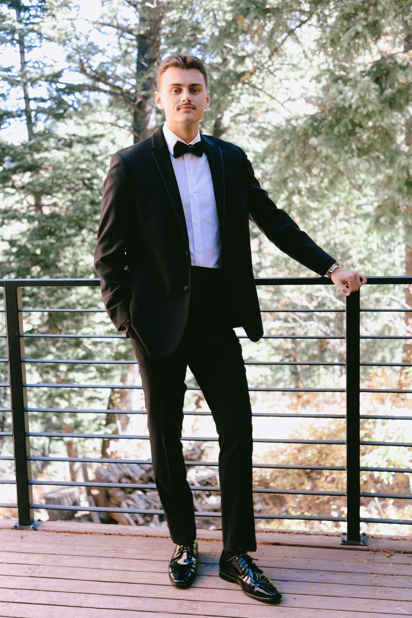 Kyle in his black tuxedo on the deck of a timber house, conifers behind him
