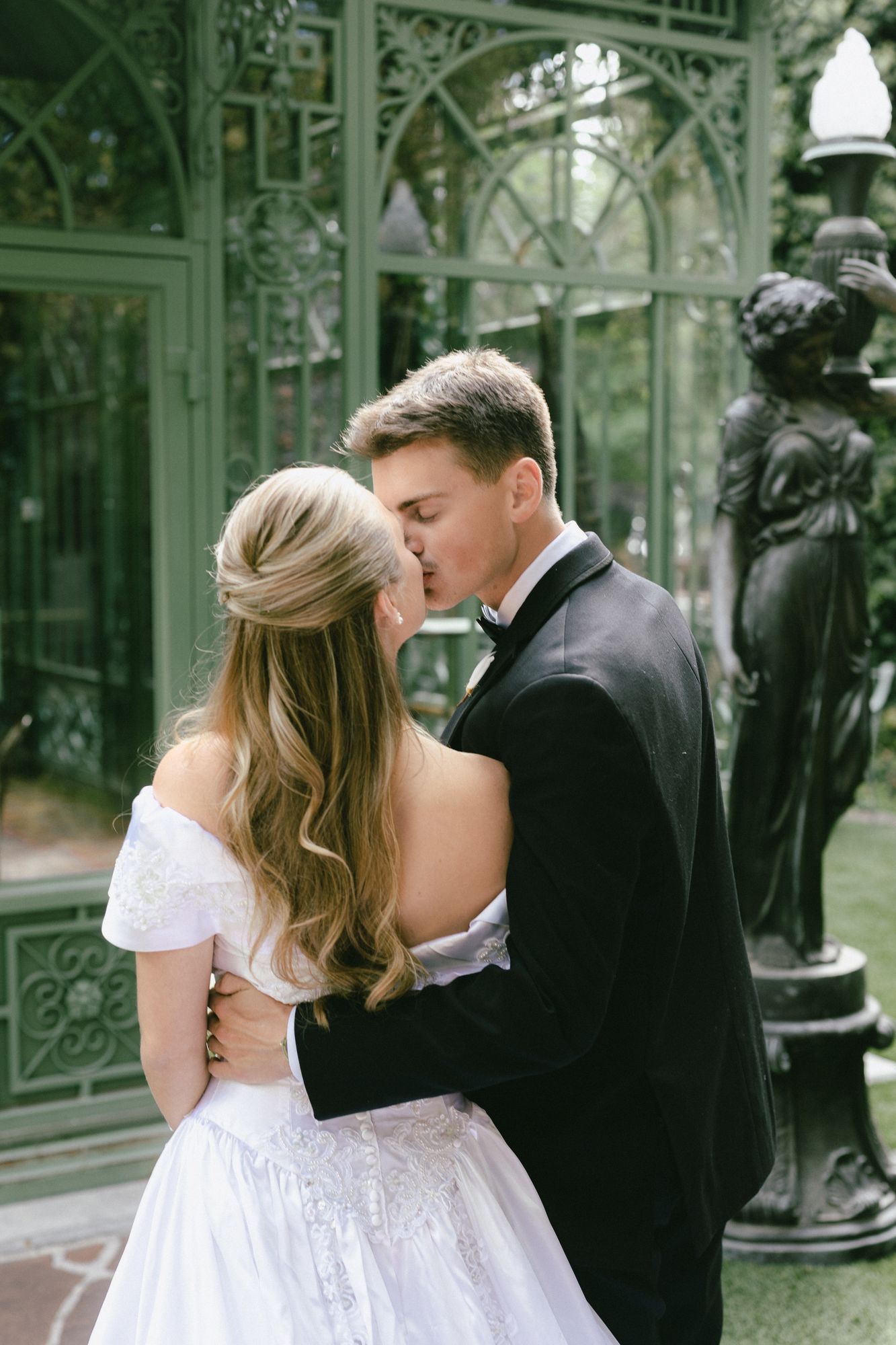 Kyle and Marita kissing in front of the green ironwork conservatory and its statuary at Bellissimo Gardens