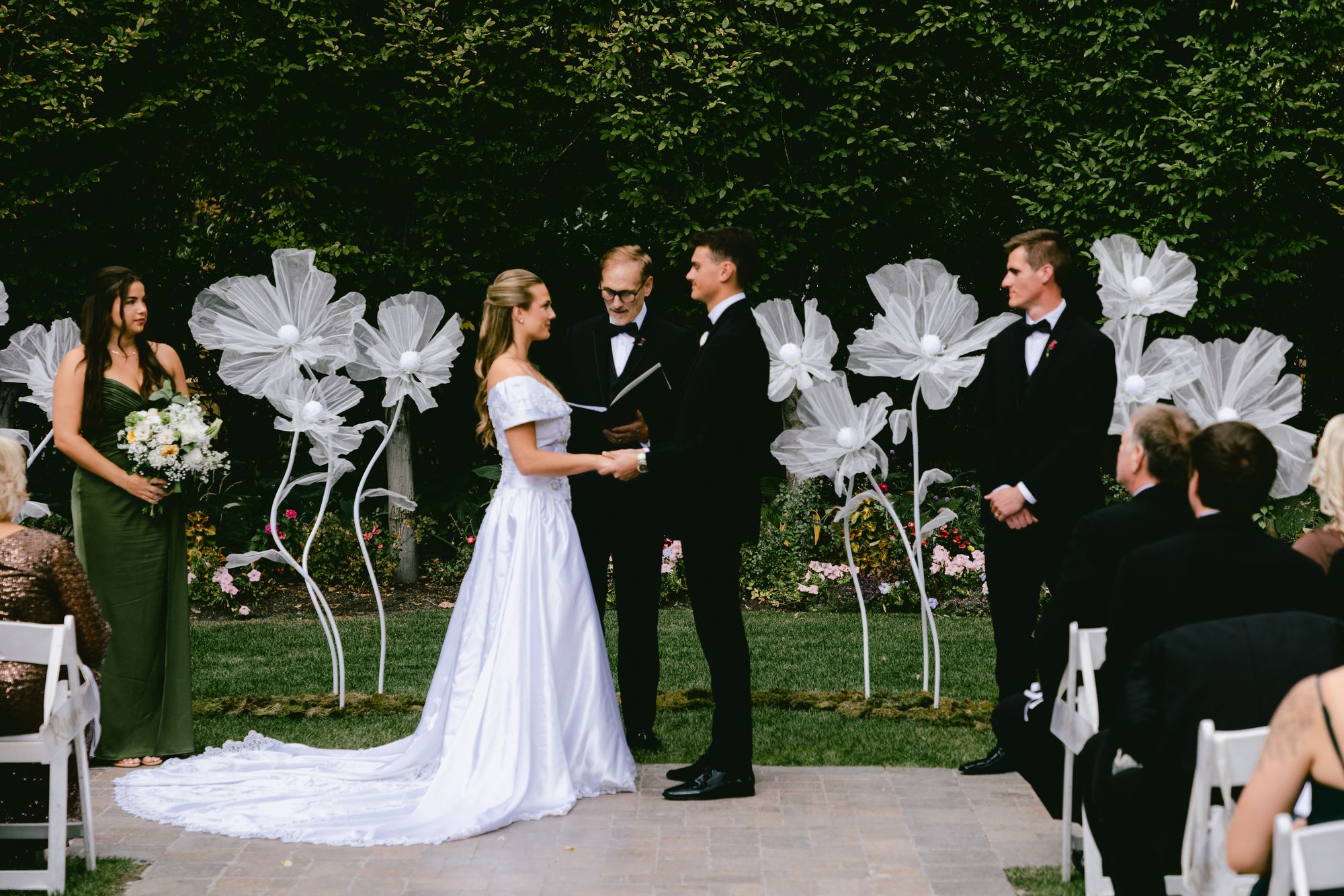 Kyle and Marita holding hands during their ceremony at Bellissimo Gardens, oversized white paper flowers on either side of them