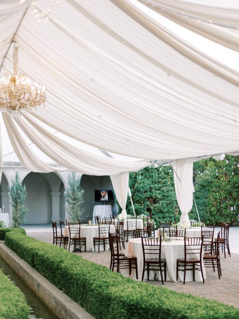 Experience enchantment with an elegant summer wedding tent in Loggia Gardens. Surrounded by lush greenery and blooming flowers, this dreamy backdrop promises a romantic celebration of love. The luxurious ambiance is heightened by sparkling lights and soft drapes, while elegant table settings and stunning floral arrangements ensure that every detail is perfect. Whether exchanging vows or dancing the night away, this exquisite setup will make your special day truly unforgettable.